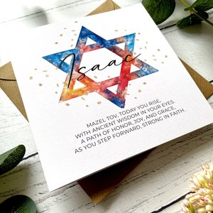 May include: A white greeting card featuring the name "Isaac" in cursive, inside a watercolour Star of David in blue, orange, and red. The card includes the text "Mazel Tov..." and is on a white wooden surface.