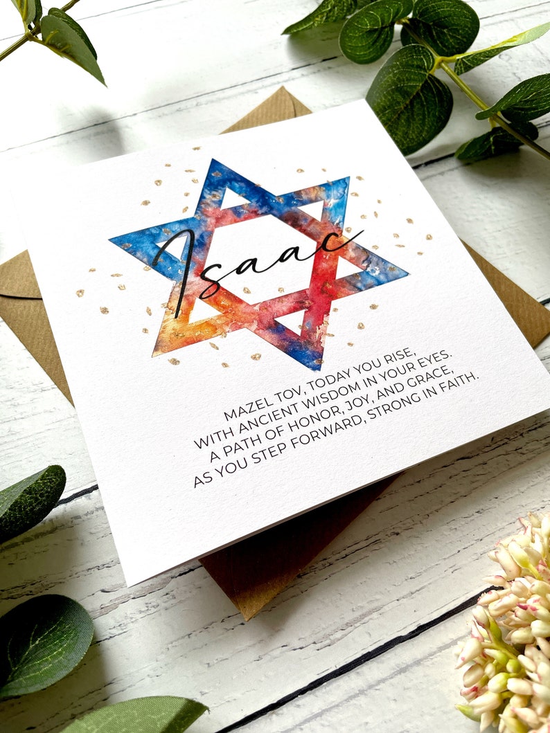 May include: A white greeting card featuring the name "Isaac" in cursive, inside a watercolour Star of David in blue, orange, and red. The card includes the text "Mazel Tov..." and is on a white wooden surface.
