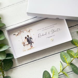 May include: A white gift box containing a wedding card. The card displays a watercolour illustration of a couple on a swing, adorned with floral details. It includes the names "Rachael & Stuart" and the date "2nd September 2025".