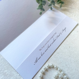 May include: A white greeting card with black printed text that reads, We hope you have a magical day! All our love and best wishes Dad & Angie. The card is accented with a sprig of green eucalyptus leaves and a strand of white faux pearls.