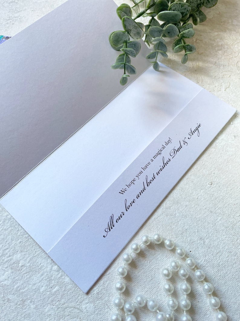 May include: A white greeting card with black printed text that reads, We hope you have a magical day! All our love and best wishes Dad & Angie. The card is accented with a sprig of green eucalyptus leaves and a strand of white faux pearls.