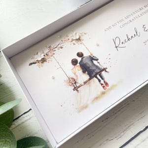 May include: A white gift box containing a card with a watercolor illustration of a couple on a swing under a floral arch. The text reads AND SO THE ADVENTURE BEGINS CONGRATULATIONS Rachael & 2nd. The card is decorated with small white pearl accents.