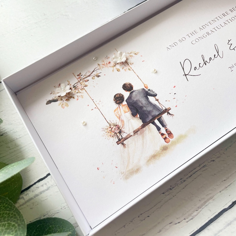 May include: A white gift box containing a card with a watercolor illustration of a couple on a swing under a floral arch. The text reads AND SO THE ADVENTURE BEGINS CONGRATULATIONS Rachael & 2nd. The card is decorated with small white pearl accents.