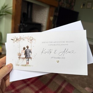 May include: A rectangular white wedding card featuring an illustration of a couple on a swing. Text reads: AND SO THE ADVENTURE BEGINS... CONGRATULATIONS, Kristy & Alex, 14th November 2026. A small heart embellishment is below the date.
