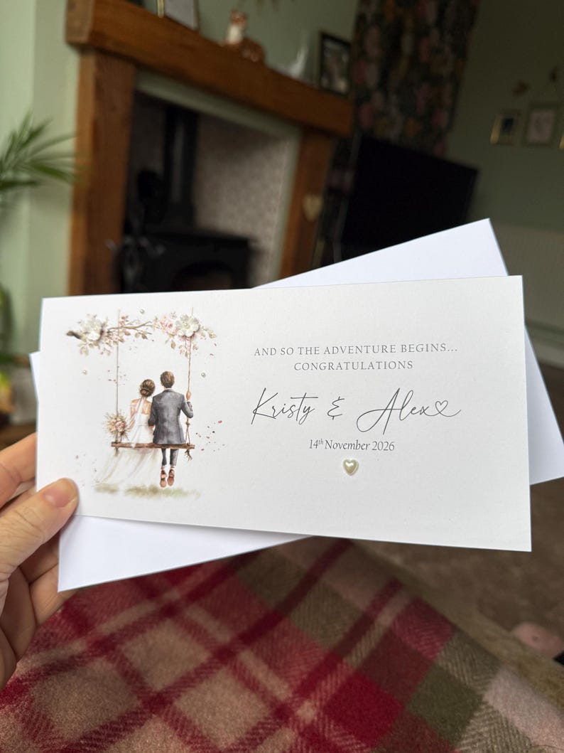 May include: A rectangular white wedding card featuring an illustration of a couple on a swing. Text reads: AND SO THE ADVENTURE BEGINS... CONGRATULATIONS, Kristy & Alex, 14th November 2026. A small heart embellishment is below the date.