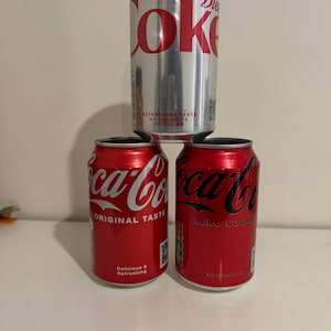 May include: A stack of three aluminium soda cans. The top can is silver with red Diet Coke branding. The bottom left can is red with white Coca-Cola Original Taste text. The bottom right can is red with black Coca-Cola Zero Sugar branding.