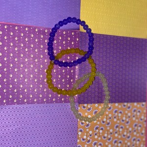 May include: Three silicone bracelets in different colours: purple, yellow and clear. The bracelets are stacked on top of each other and are lying on a multi-coloured patterned surface.