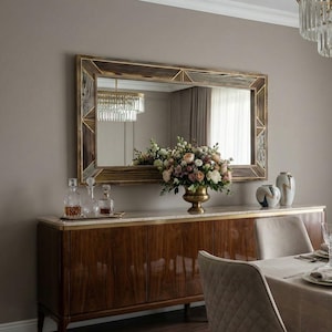 Puede incluir: Un comedor con un gran espejo rectangular en un marco de madera decorativo sobre un aparador de madera. El aparador tiene una tapa de mármol y contiene un arreglo floral, jarrones y decantadores. Una mesa de comedor con cubiertos está en primer plano.