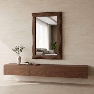 May include: A rectangular, dark wood-framed mirror with a wavy edge hangs above a matching floating cabinet. The cabinet has three drawers. A vase with greenery and a stack of books sit on the cabinet. The room has a neutral color palette.