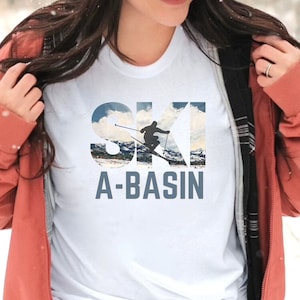 May include: White t-shirt with the words "SKI A-BASIN" in a blue-gray font. The word "SKI" has a mountain and sky scene as the background. A skier is in the middle of the word "SKI". The person is wearing a red jacket.