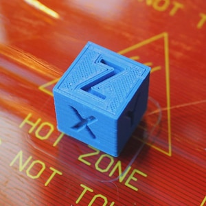 Puede incluir: Un pequeño cubo azul impreso en 3D con las letras "X" y "Z" recortadas en lados adyacentes. El cubo está sobre una superficie roja con texto amarillo "HOT ZONE NOT TO". El cubo es una forma geométrica sólida.