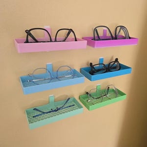 Puede incluir: Colección de gafas en bandejas de pared coloridas. Las bandejas son rosas, moradas, azules y verdes. Cada bandeja contiene un par de gafas, con monturas negras y plateadas.