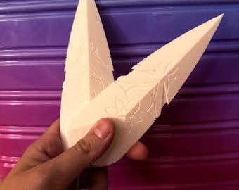 May include: Two off-white, feather-shaped decorative pieces with floral and leaf patterns. The pieces have a pointed tip and are held against a background that transitions from purple to pink. A small, orange tassel hangs from the bottom.