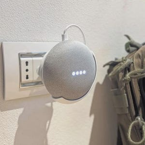 Puede incluir: Un altavoz inteligente gris claro con un cable de alimentación blanco está enchufado a una toma de corriente. El altavoz tiene cuatro puntos blancos iluminados. Una mochila de color canela es parcialmente visible en el fondo.
