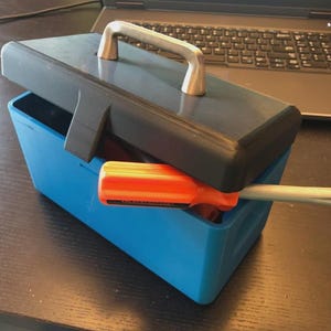 Puede incluir: Una caja de herramientas azul con tapa negra y asa plateada. Un destornillador naranja y plateado está parcialmente dentro de la caja de herramientas. La caja de herramientas está sobre una superficie oscura.