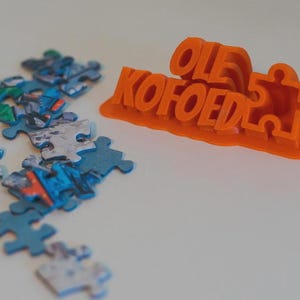 May include: An orange puzzle-shaped object with the name "OLE KOFOED" in raised letters. Scattered around are several puzzle pieces in shades of blue, green, and gray, suggesting a puzzle-themed item.