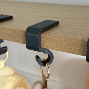 May include: Black metal shelf hooks with a curved design, attached to a light wood shelf. The hooks are holding gold-colored keychains and plush toys. The design is modern and minimalist.