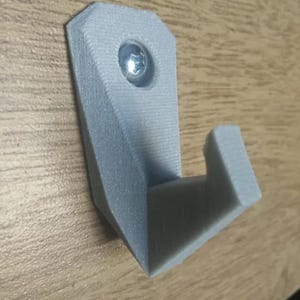 May include: A light gray, 3D-printed wall hook with a screw. The hook has a geometric design with a rectangular base and a curved hook. The screw is silver and is used to attach the hook to a wall or other surface.