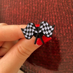 Può includere: Uno spillo a forma di bandiera a scacchi nera e bianca. Lo spillo è in metallo e ha un retro nero. Le bandiere sono incrociate e lo spillo è attaccato a un retro rosso.