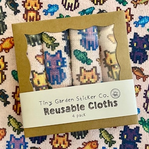 Op de afbeelding: Een set van vier herbruikbare doeken met pixelkat- en visontwerpen. De doeken zijn zichtbaar door een venster in de kartonnen doos, met de tekst "Tiny Garden Sticker Co. Reusable Cloths 4 pack."