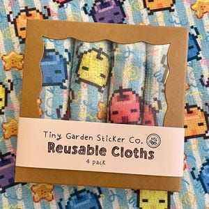 May include: A brown cardboard box with a scalloped window, containing four rolled reusable cloths. The cloths have a light blue background with pixelated characters. The box has a white label that reads "Tiny Garden Sticker Co. Reusable Cloths 4 pack."