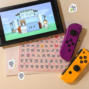 May include: A purple and yellow Nintendo Switch Joy-Con controller set with a pink and white sticker sheet featuring a cartoon cat design. The sticker sheet has a variety of cat expressions and poses. The Switch console is in the background, displaying a video game.