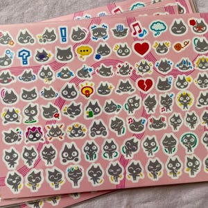 May include: A sheet of stickers featuring black cats with different expressions and actions. The stickers are on a pink background with black and white stripes.