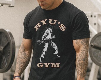 May include: Black t-shirt with the text "RYU'S GYM" in a tan color. The shirt features a graphic of a person lifting weights. The person in the graphic is wearing a karate gi. The shirt is made of a soft material.