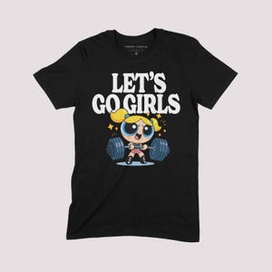 K&ouml;nnte beinhalten: Schwarzes T-Shirt mit dem wei&szlig;en Schriftzug "LET'S GO GIRLS". Das Design zeigt eine Zeichentrickfigur, die Gewichte hebt. Die Figur hat blonde Haare und tr&auml;gt ein rosa Outfit. Das T-Shirt ist von der Marke "Trippy Turtle".