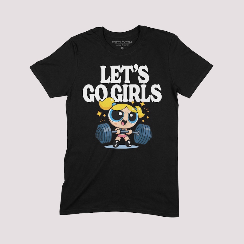 K&ouml;nnte beinhalten: Schwarzes T-Shirt mit dem wei&szlig;en Schriftzug "LET'S GO GIRLS". Das Design zeigt eine Zeichentrickfigur, die Gewichte hebt. Die Figur hat blonde Haare und tr&auml;gt ein rosa Outfit. Das T-Shirt ist von der Marke "Trippy Turtle".