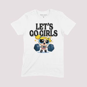 K&ouml;nnte beinhalten: Wei&szlig;es T-Shirt mit einer Cartoon-Illustration einer Figur, die Gewichte hebt, und dem Text "LET'S GO GIRLS". Das Design zeigt eine blonde Figur mit blauen Augen und eine Langhantel mit blauen Gewichten. Das Shirt hat einen Rundhalsausschnitt.