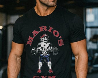 May include: Black t-shirt featuring a muscular cartoon Mario character lifting weights, with the text "Mario's Gym" in red. The design includes a red hat, blue shorts, and red shoes. The shirt is short-sleeved.