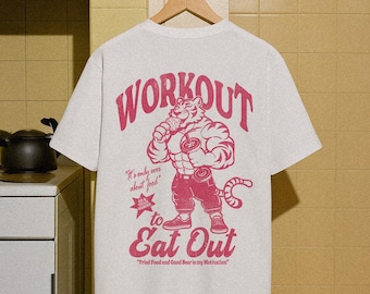 May include: A light gray t-shirt with a red graphic of a muscular tiger holding a drink and weights. The text "WORKOUT" arches across the top, with "to Eat Out" below the tiger. Additional text reads "It's only ever about food" and "Fried Food and Good Beer is my Motivation."