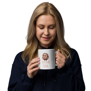 Puede incluir: Taza de cerámica blanca con la cara de una mujer y el texto "NAME 'EM." impreso en color melocotón.