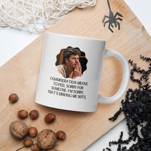 Può includere: Tazza in ceramica bianca con manico, con una foto di un uomo e il testo: "COMMISERATION MEANS TO FEEL SORRY FOR SOMEONE. I'M SORRY, THAT'S DRIVING ME NUTS." La tazza è su una superficie di legno con noci.