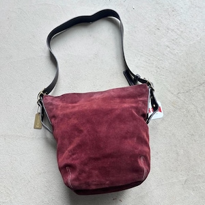 May include: A burgundy suede shoulder bag with a black leather strap and a gold buckle. The bag has a top zipper closure and a small pocket on the front.