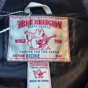 May include: Close-up of a black jacket's interior with two tags. The larger tag reads "TRUE RELIGION BRAND JEANS" with a graphic of a Buddha playing a guitar. The smaller tag also has the brand name and "MADE IN CHINA" with a size "L".