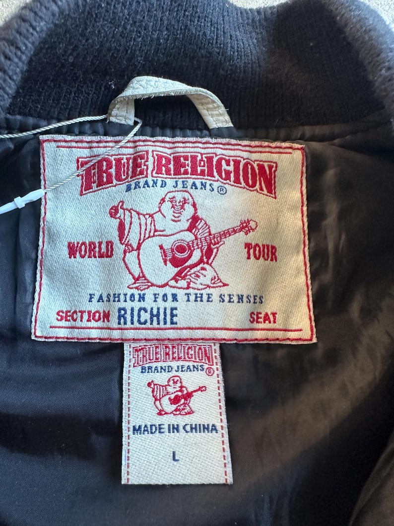 May include: Close-up of a black jacket's interior with two tags. The larger tag reads "TRUE RELIGION BRAND JEANS" with a graphic of a Buddha playing a guitar. The smaller tag also has the brand name and "MADE IN CHINA" with a size "L".