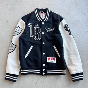 May include: Black and cream varsity jacket with white accents. Features the text "TRBJ" and "True Religion" on the front. The sleeves are cream-colored with patches. A classic, stylish outerwear piece.