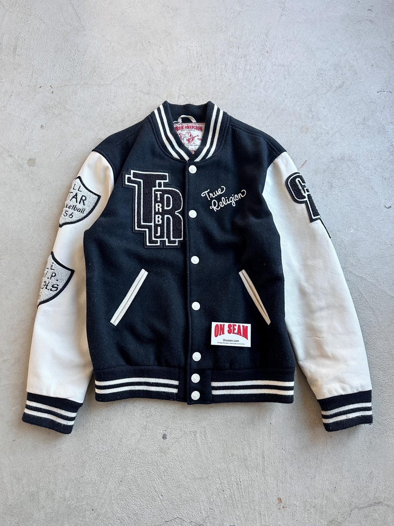May include: Black and cream varsity jacket with white accents. Features the text "TRBJ" and "True Religion" on the front. The sleeves are cream-colored with patches. A classic, stylish outerwear piece.