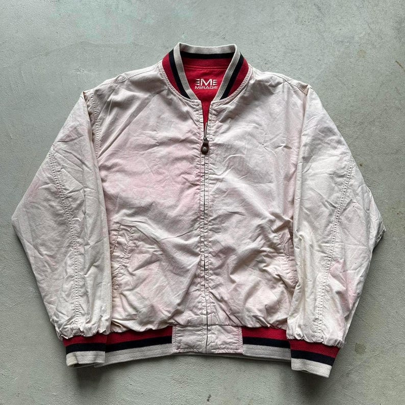 May include: A white bomber jacket with a red and black striped collar and cuffs. The jacket has a zipper closure and the text "EME MIRAGE" is printed on the front.