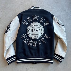 May include: Black varsity jacket with white leather sleeves. The back features a circular emblem with "HAWKS", "BROOKLYN CHAMPS", "VARSITY", and "1956" text. The jacket has a striped collar, cuffs, and hem.