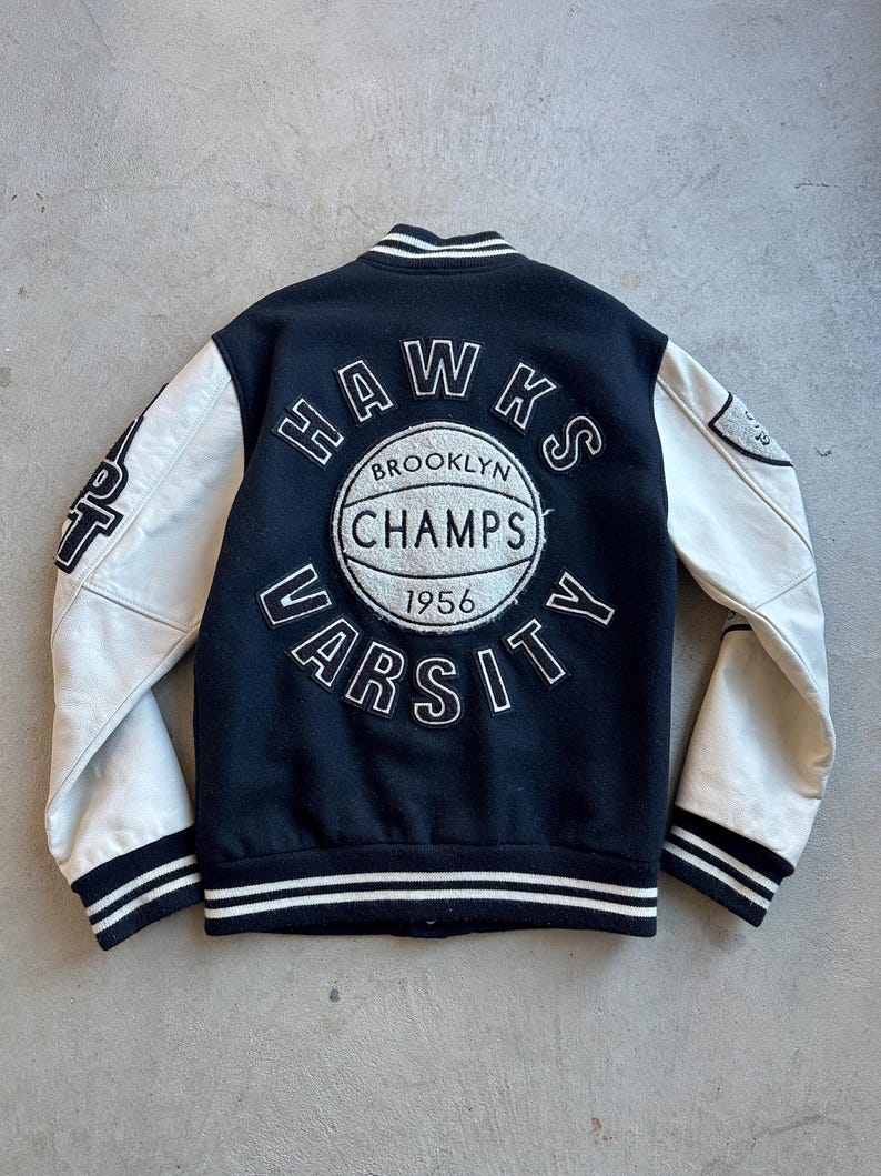May include: Black varsity jacket with white leather sleeves. The back features a circular emblem with "HAWKS", "BROOKLYN CHAMPS", "VARSITY", and "1956" text. The jacket has a striped collar, cuffs, and hem.