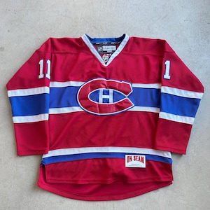 May include: A red hockey jersey with white and blue accents. The jersey features the number 11 on the left sleeve and the number 1 on the right sleeve. The team logo is a large "C" with an "H" inside. The jersey has a V-neck collar.