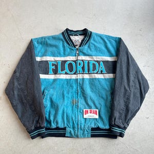 May include: A turquoise and black bomber jacket with the word "FLORIDA" across the chest. The jacket has a white stripe and black sleeves. The tag "ON SEAM" is visible on the lower right.