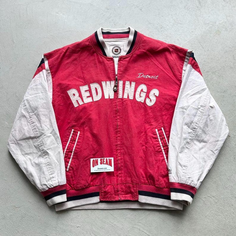 May include: A red and white baseball jacket with the words "Detroit Redwings" on the front. The jacket has a zipper closure and a ribbed collar, cuffs, and waistband.