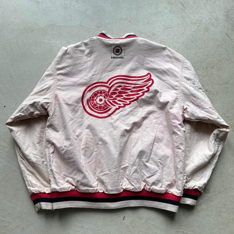 May include: A white baseball jacket with a red and white Detroit Red Wings logo on the back. The jacket has a black and red striped waistband and cuffs. The text "MIRAGE" is printed on the back of the jacket.