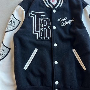 May include: Black and white varsity jacket with white sleeves. Features the text "True Religion" in script and a large "TRBJ" patch. Additional patches on the sleeves include text and a shield design. The jacket has a button-up front and white trim.