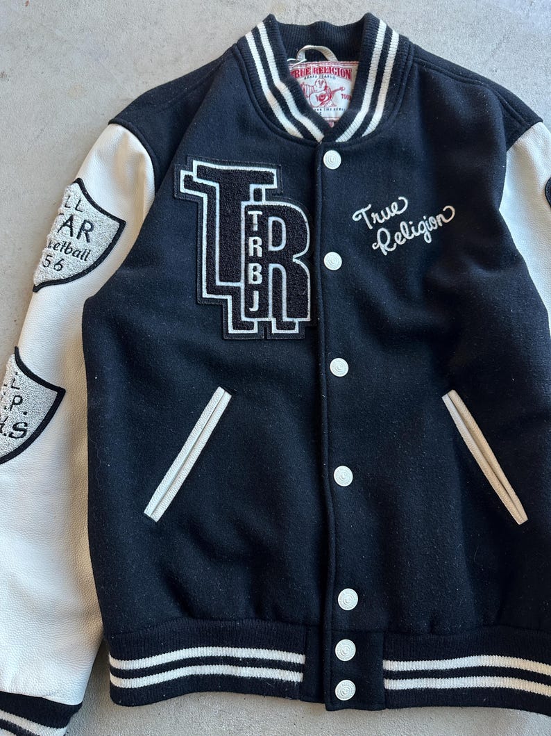 May include: Black and white varsity jacket with white sleeves. Features the text "True Religion" in script and a large "TRBJ" patch. Additional patches on the sleeves include text and a shield design. The jacket has a button-up front and white trim.
