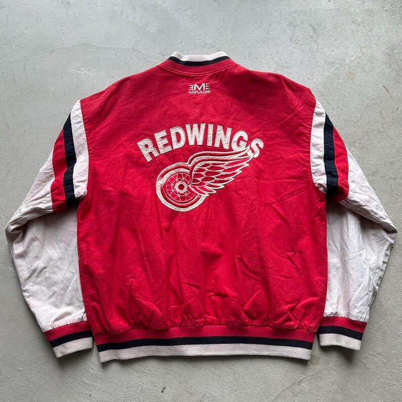 May include: Red and white baseball jacket with the Detroit Red Wings logo on the back. The logo is white with a red wing and a wheel. The jacket has black and white stripes on the sleeves.